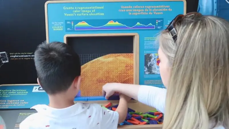 This Image Shows an Interactive Exhibit at Science Central in Fort Wayne, Where a Child and An Adult Are Using Colorful Wooden Pieces to Create a Representational Color Image of Venus's Surface Elevation