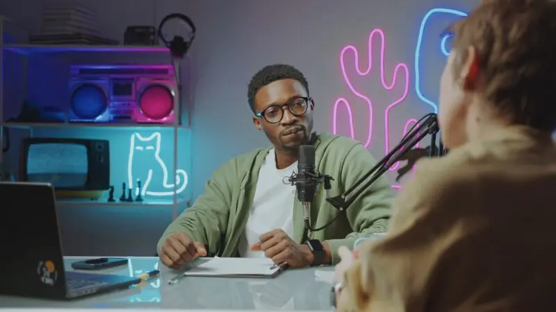 A man wearing glasses and headphones sitting at a table in a neon-lit room, speaking into a microphone