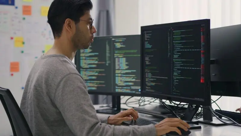 A software developer in a gray sweater and glasses sits at a workstation with multiple monitors displaying complex lines of code