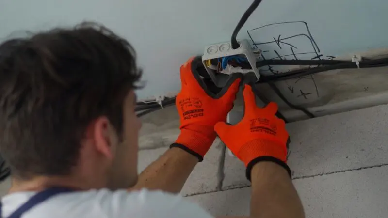 A male electrician wearing bright orange safety gloves installs electrical wiring in a junction box on a ceiling