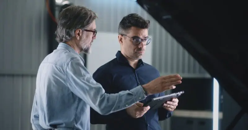 Two male engineers stand in a well-lit industrial setting, discussing a project while reviewing a tablet