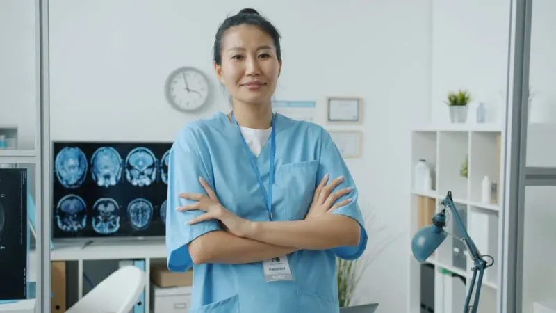 A female nurse in blue scrubs stands confidently in a well-lit medical office with arms crossed