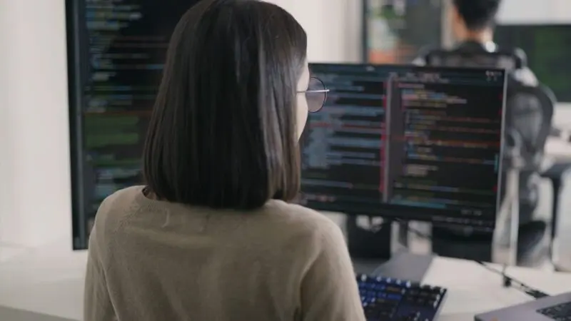 A female software engineer with shoulder-length dark hair sits at a desk, reviewing lines of code on dual monitors.